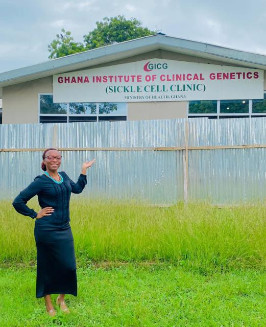 A person stands smiling and gesturing towards the Ghana Institute of Clinical Genetics (Sickle Cell Clinic) building. 