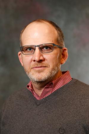 Portrait of a person wearing glasses, a red checkered shirt, and standing against a mottled gray background.