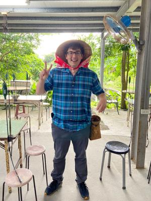 An individual wearing a plaid shirt and jeans stands in a workshop surrounded by metal sculptures, holding up his fingers in a peace sign.
