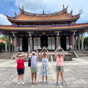 Four people pose with their arms to form O-H-I-O in front of the ornate Confucius Temple under a blue sky.