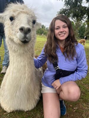 Person smiling while seated next to a white alpaca in a grassy field.