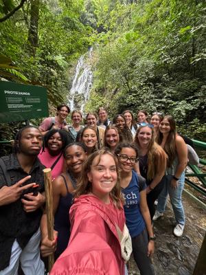 Group of students taking a selfie in front of a waterfall in a lush green forest.