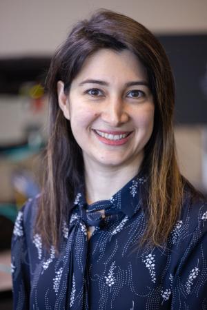 Portrait of a smiling individual in a dark blouse with a light pattern, standing in a laboratory setting.