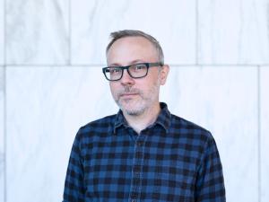 Portrait of an individual wearing glasses and a plaid shirt, standing against a marble background.