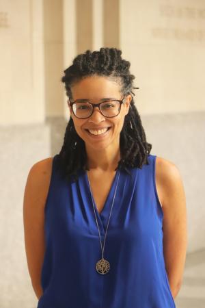 Person with glasses smiling, wearing a blue top and a pendant necklace, standing in a hallway.