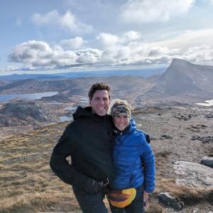 Two individuals posing on a mountain top with scenic views of lakes and rolling hills in the background.