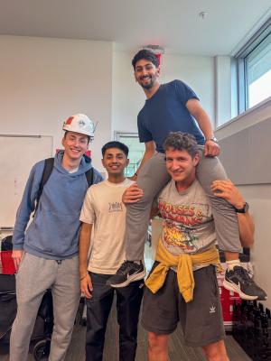 Four smiling individuals posing for a photo in a classroom, with one person sitting on the shoulders of another, all wearing casual attire.