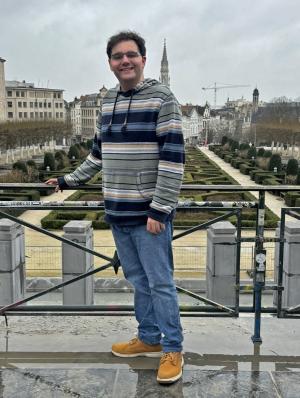 A person in a striped hoodie, blue jeans, and yellow boots standing on a balcony overlooking a formal garden with trimmed hedges and pathways, with a cityscape and a tall spire in the background.