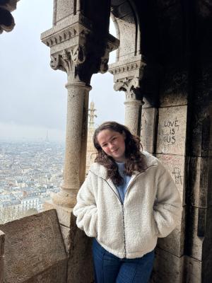 A person smiling at the camera, standing inside a stone archway with a view of Paris and the Eiffel Tower in the background.