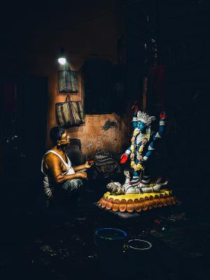 An artist painting a colorful statue of the Hindu deity Krishna in a dimly lit workshop.