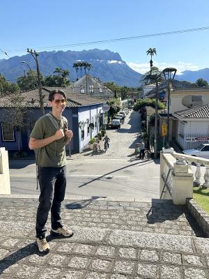 Person standing on stone steps in a quaint town, smiling towards the camera with a scenic mountain range in the background.