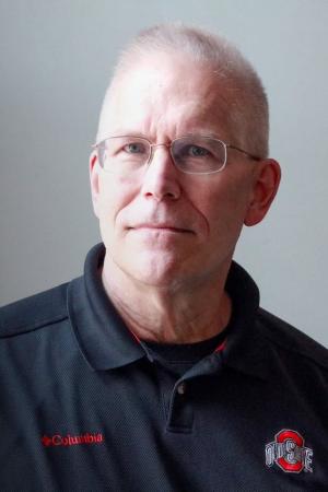 Person wearing glasses and a black Columbia polo shirt with an Ohio State logo, standing against a light gray background.